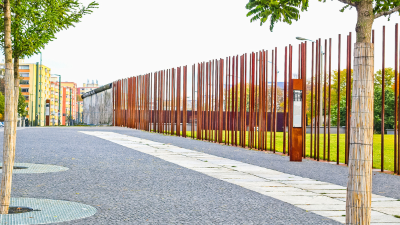 Ein Abschnitt der Berliner Mauer in der Bernauer Straße mit historischen Wachtürmen und Informationsschildern, die die Geschichte der Mauer und die Teilung Berlins dokumentieren. Im Hintergrund sind grüne Bäume und Spazierwege sichtbar, die heute an die bewegte Vergangenheit erinnern.